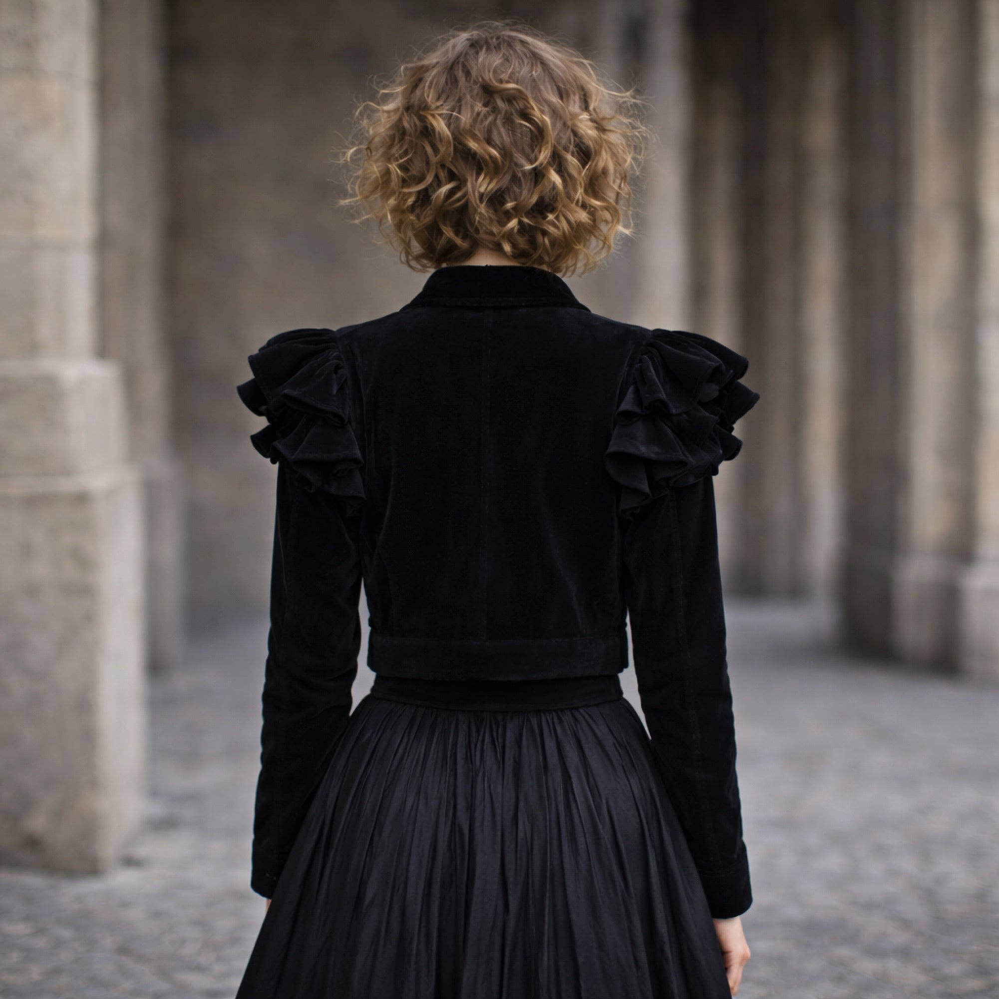 Person wearing a black velvet jacket with ruffled shoulders in an architectural setting