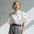 Woman wearing a white blouse with ruffled sleeves and gray pants against a neutral background